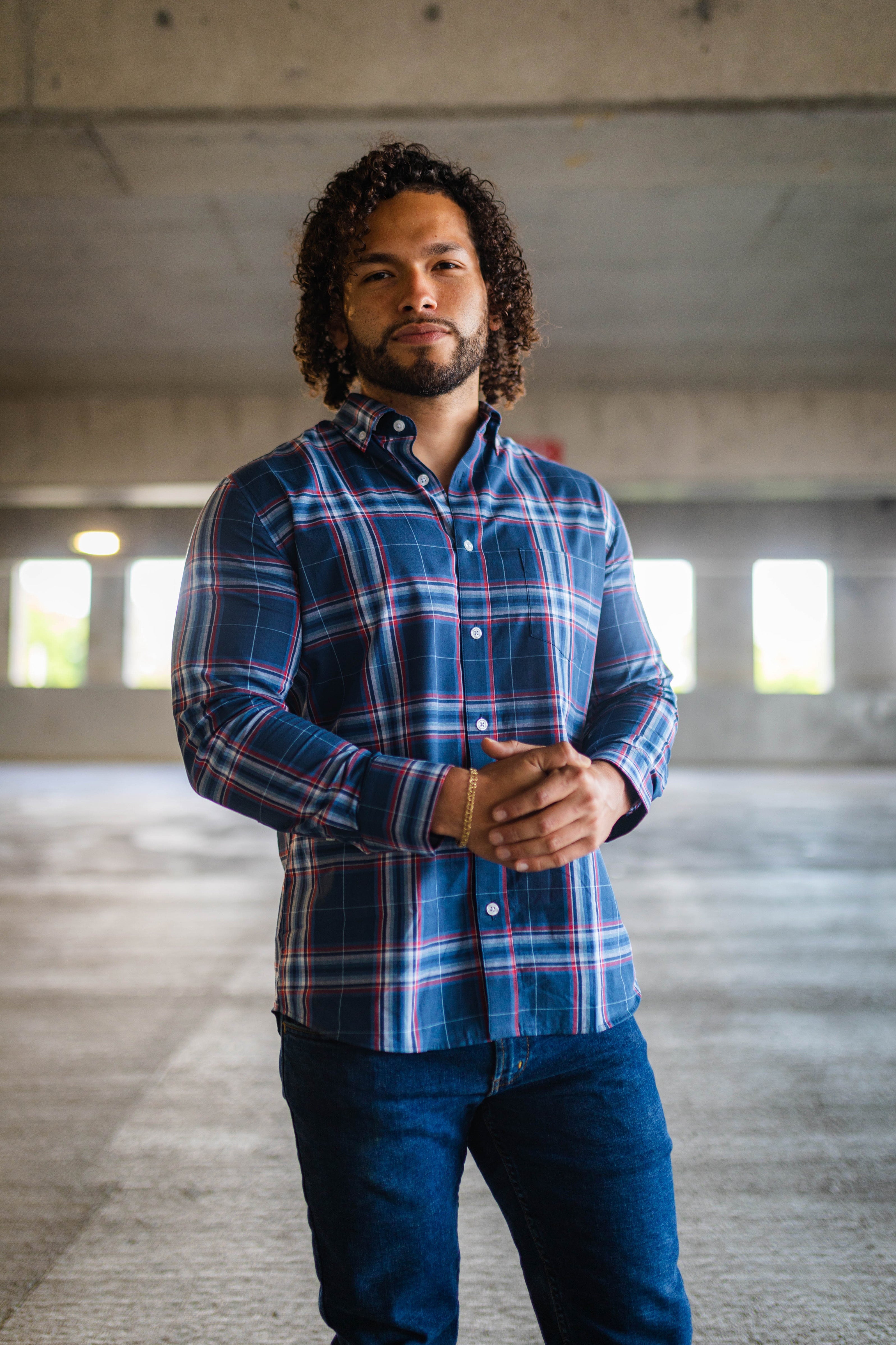 A man with curly hair and a beard is standing in a parking garage. He is wearing a blue and red plaid button-up shirt and dark blue jeans, with his hands clasped in front of him.