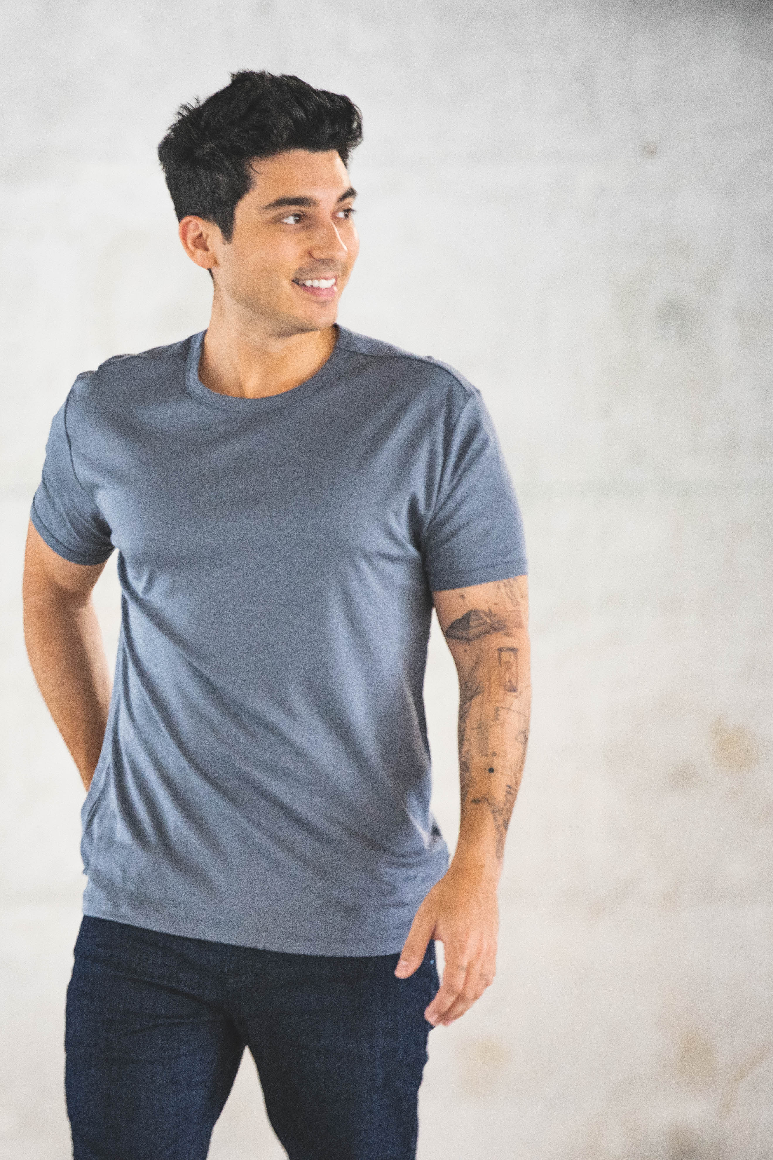 A man with short dark hair is smiling and looking to the side. He is wearing a gray t-shirt and dark blue jeans, with tattoos visible on his left arm