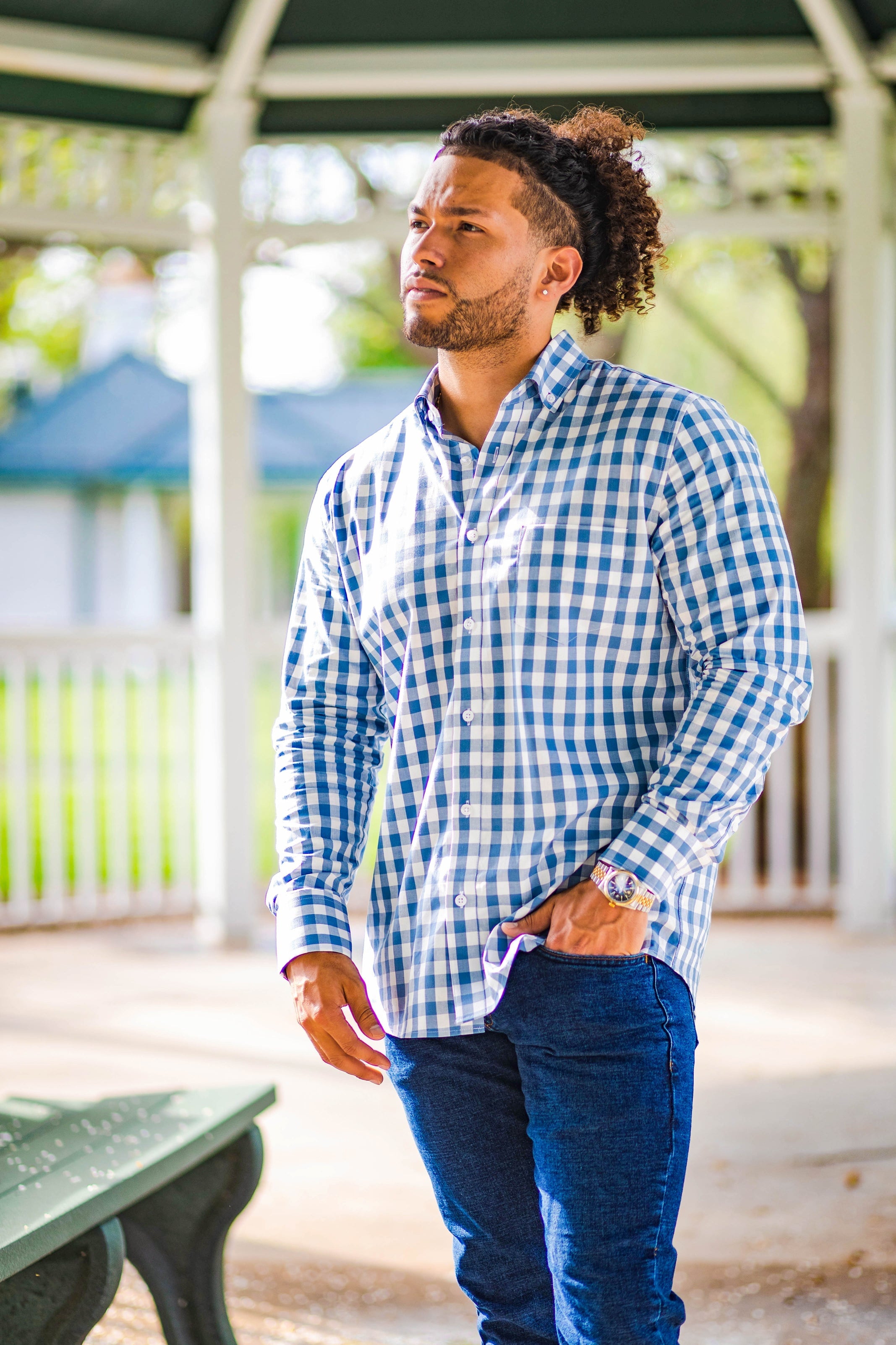 A man with curly hair is standing outdoors under a gazebo. He is wearing a blue and white checkered shirt and dark blue jeans, with his left hand in his pocket.