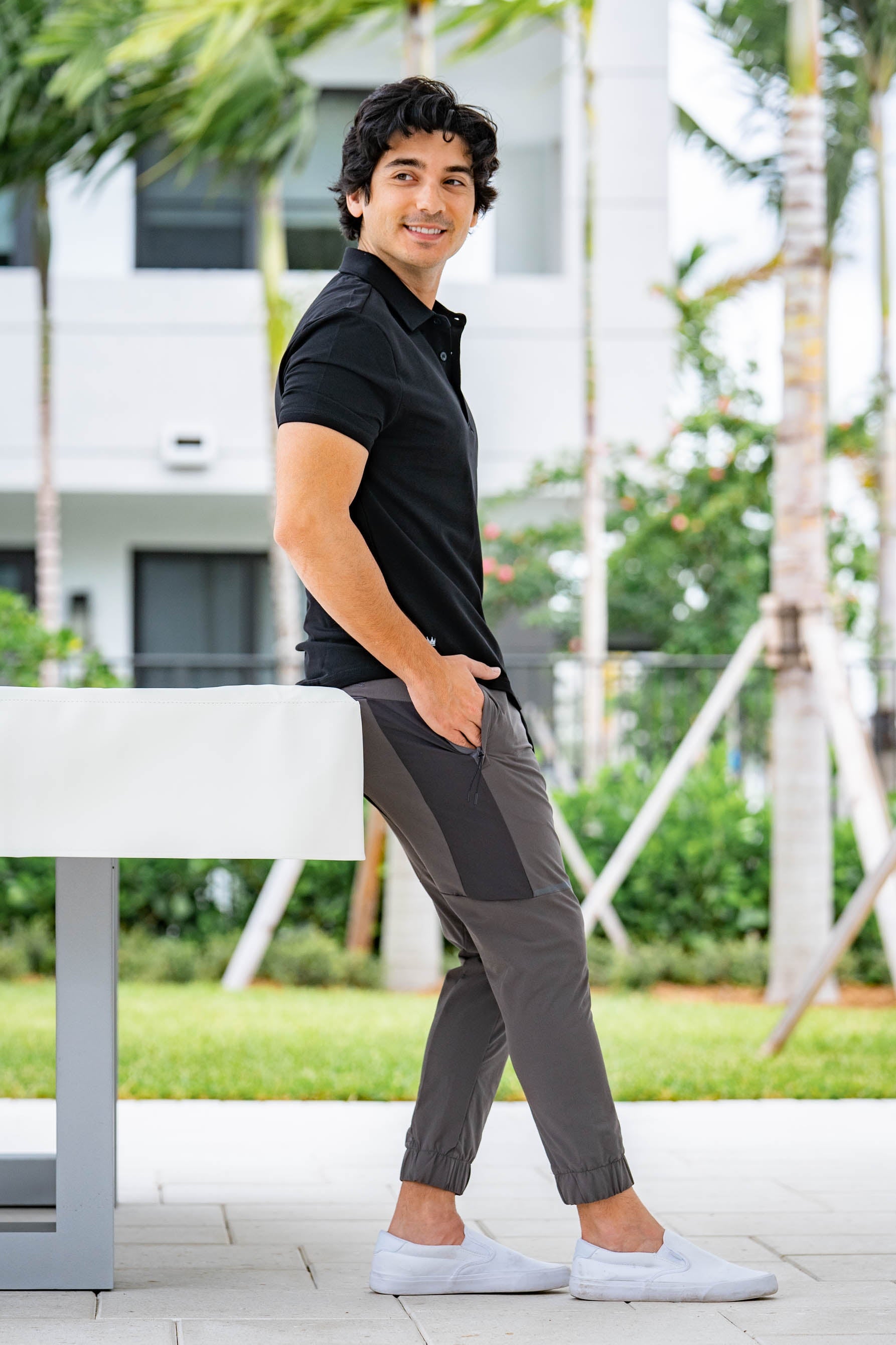 Man in a black polo shirt and gray joggers, standing outdoors with greenery in the background.