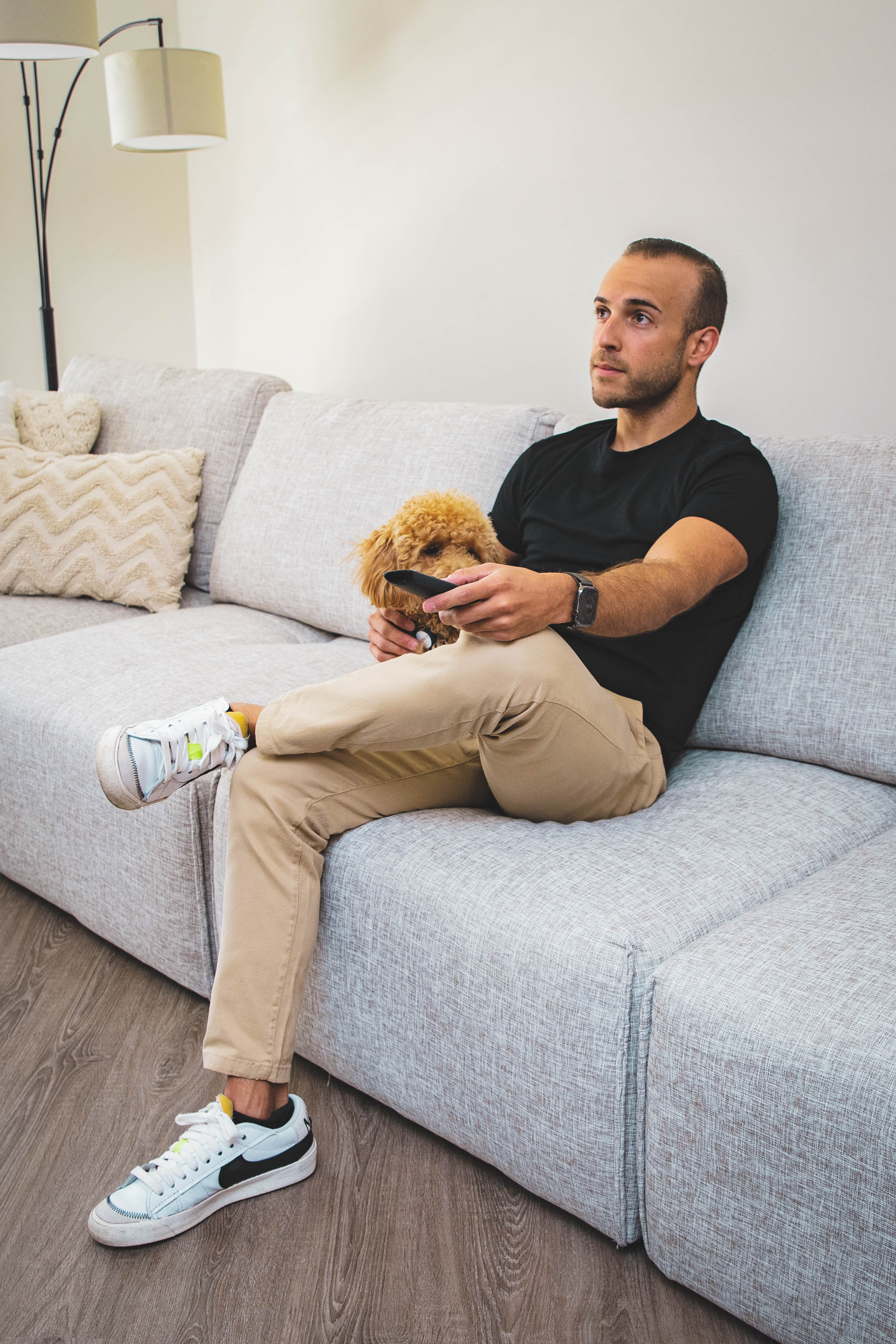 A person wearing beige pants and white sneakers, sitting on a light gray sofa, holding a small dog and looking to the side.