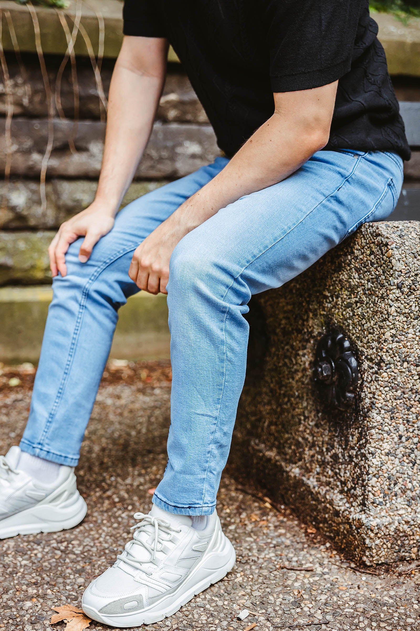 A person wearing light blue jeans and white sneakers, sitting on a stone bench with their hands resting on their knees.