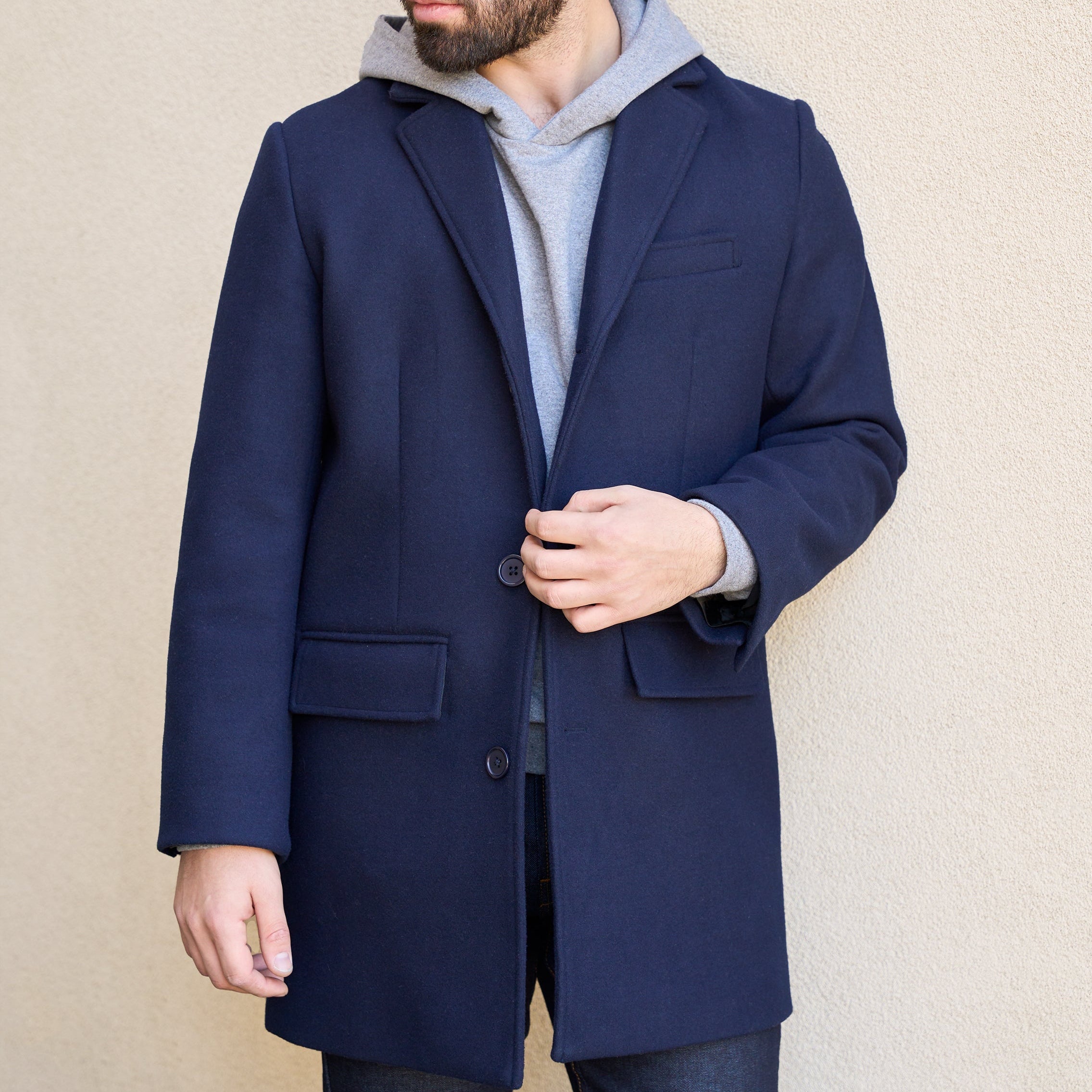 Man wearing a navy blue coat over a gray hoodie against a beige background