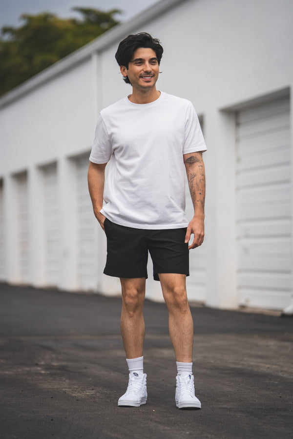 Man wearing a white t-shirt, black shorts, and white sneakers standing in front of a white building.