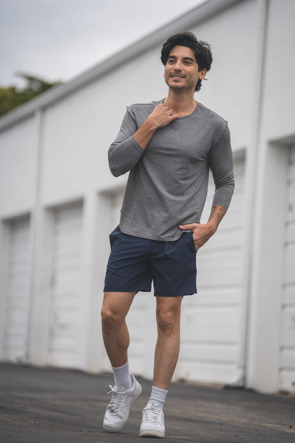 Man wearing a gray long-sleeve shirt and navy shorts standing in front of a white building.