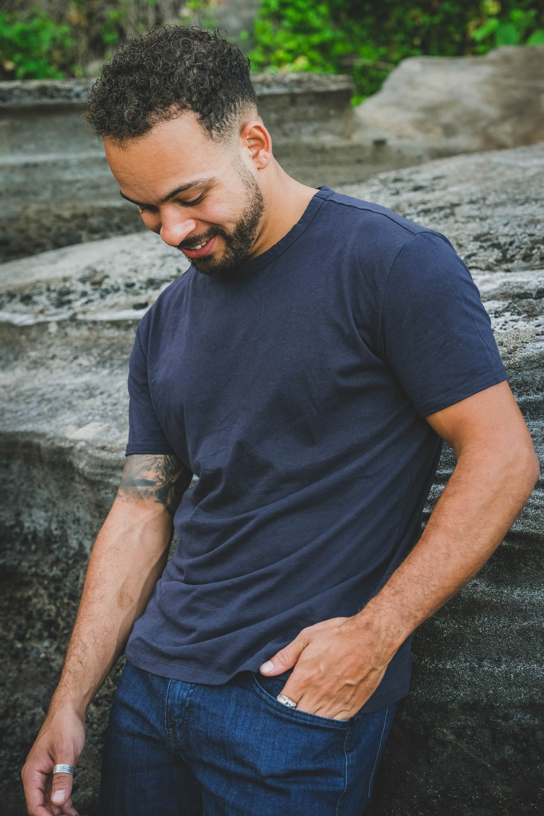 Man in a navy blue t-shirt and dark blue jeans smiling and looking down while standing by a rocky background.