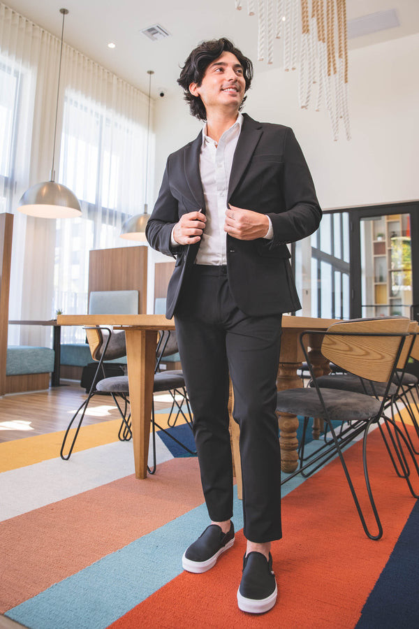 A person wearing a black stretch suit with a white shirt and black slip-on shoes, standing in a brightly lit room with modern decor and colorful carpet.