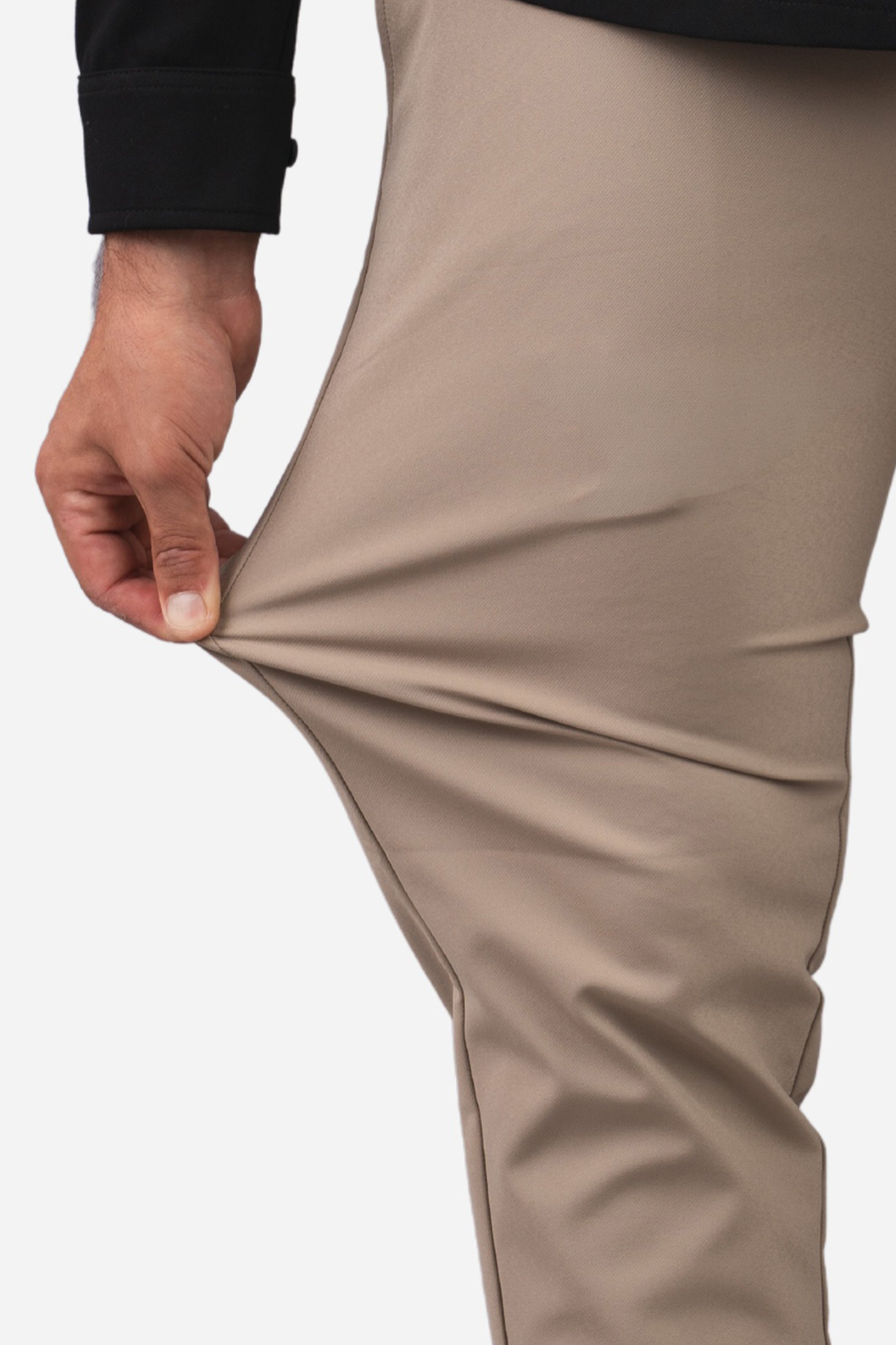 Close-up of a person wearing beige pants, pulling the fabric outward with a hand to demonstrate stretch, against a plain light background.
